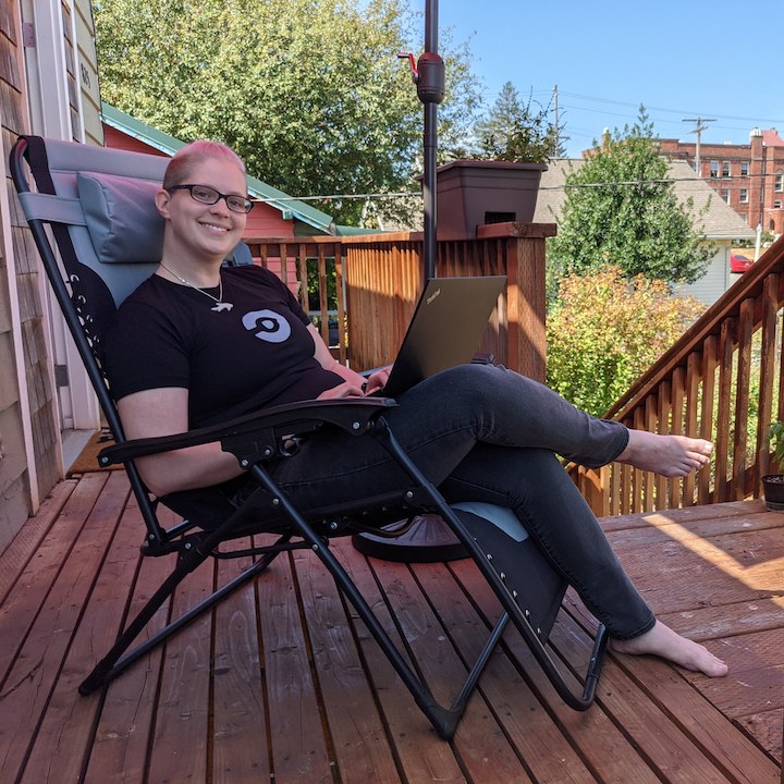 A team member smiling and sitting on a chair outside on the porch with a laptop in hand.